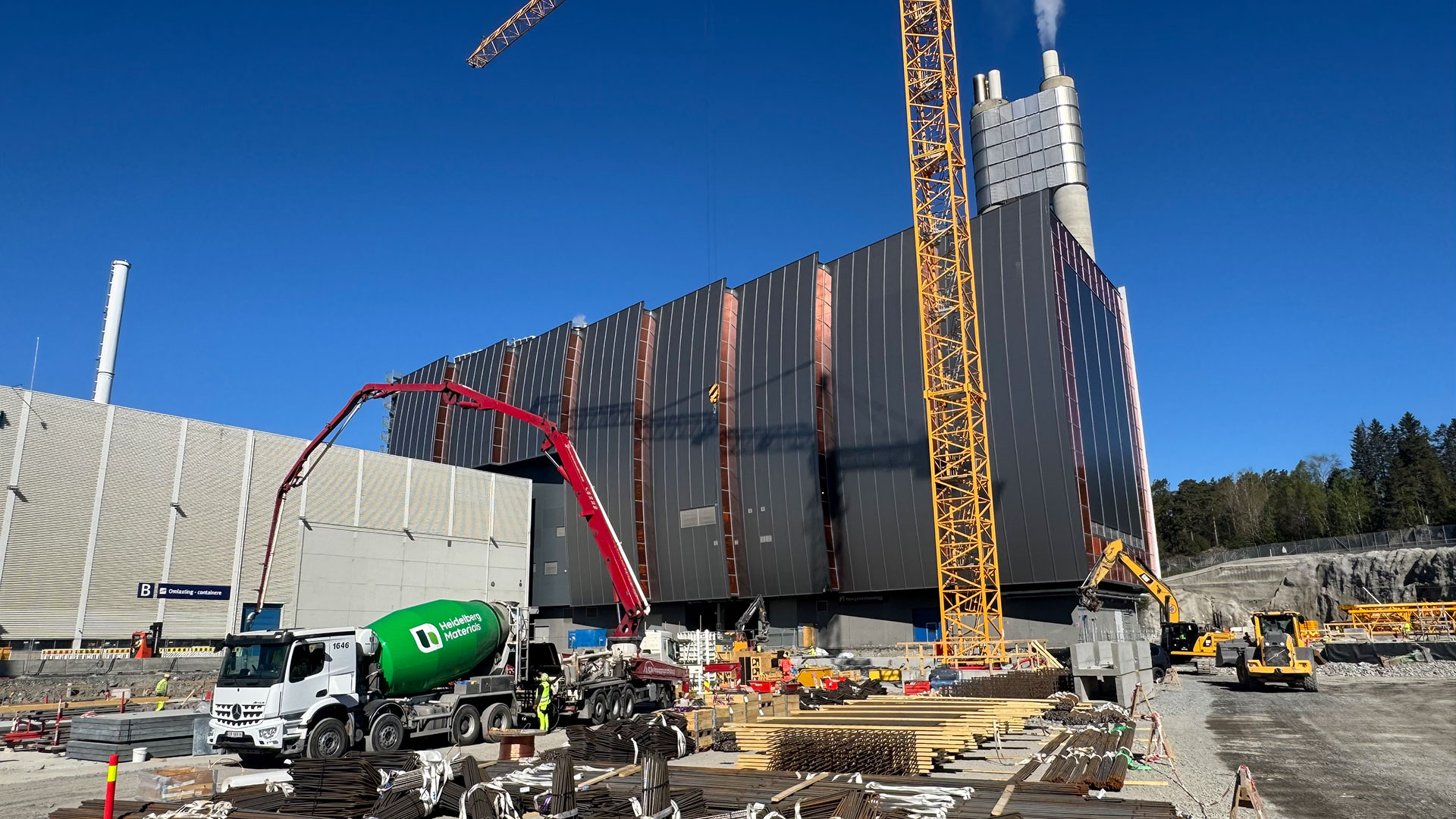 Byggeplass med stort industribygg under oppføring, inkludert kraner og betongbil.